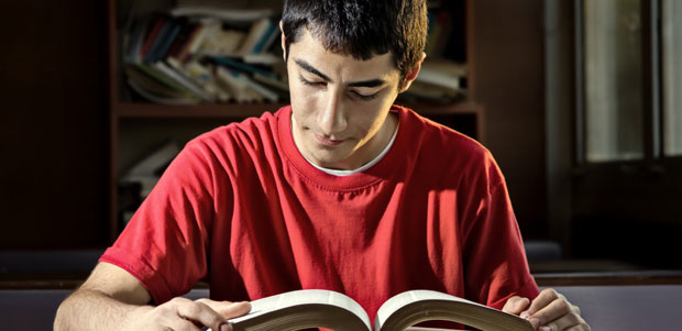 A teenage boy of Mid-Eastern descent reads a book in a darkened room.
